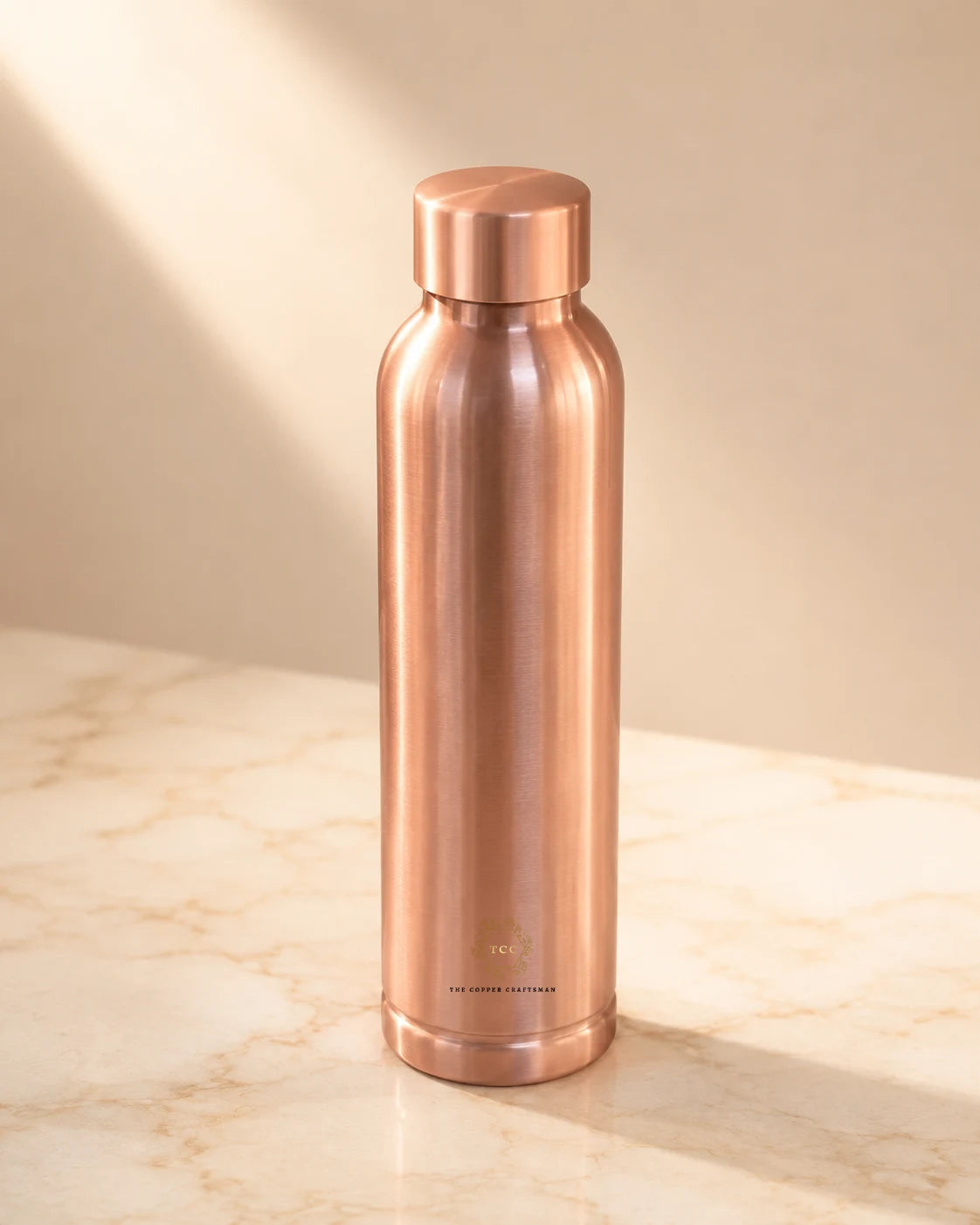 A sleek, copper water bottle (plain oreo) with a matching screw-on lid, standing on a light beige marble surface against a neutral beige background with soft diagonal shadows.