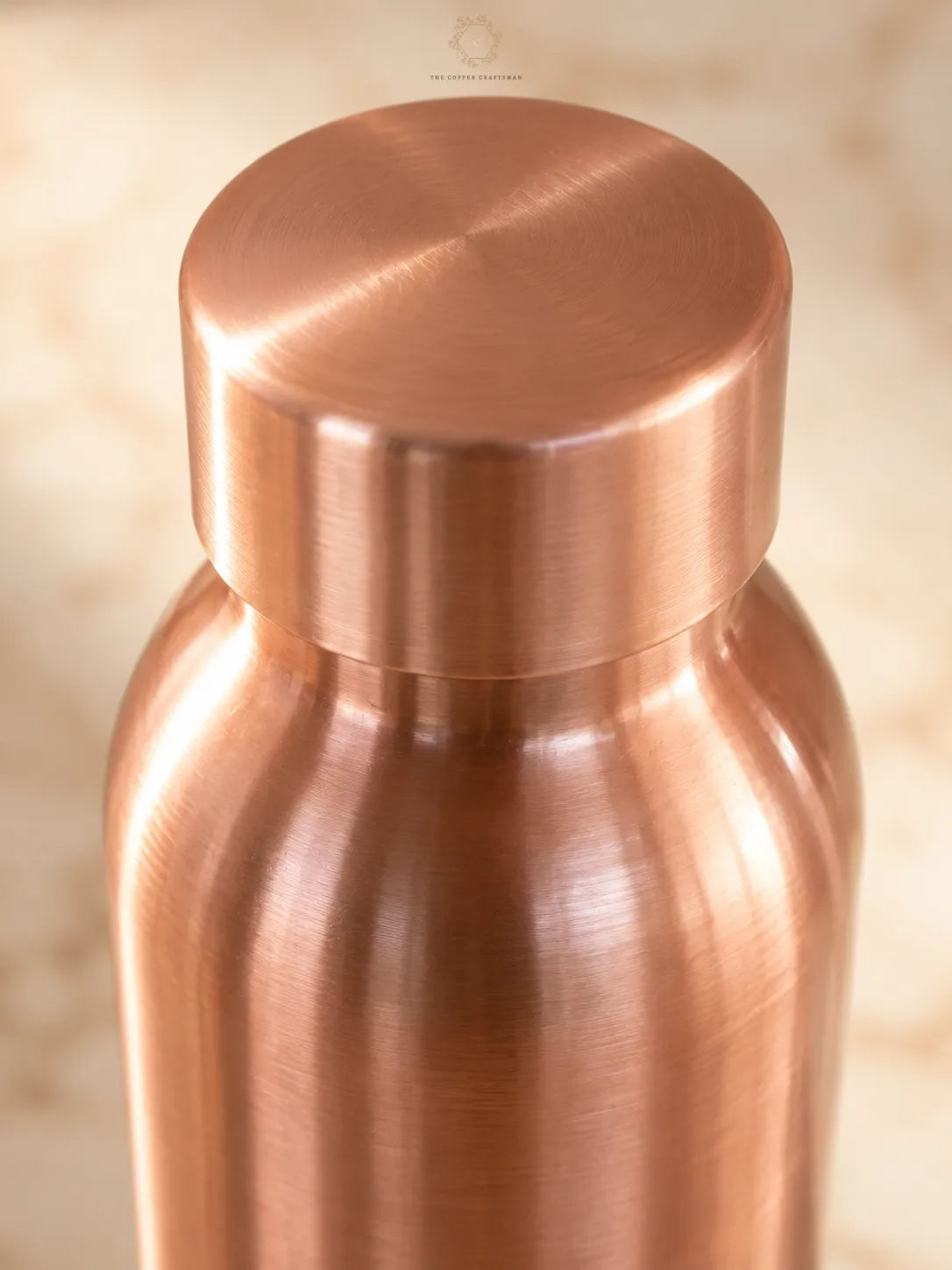 Close-up of a brushed copper bottle with a matching screw-on lid, set against a softly blurred beige background.