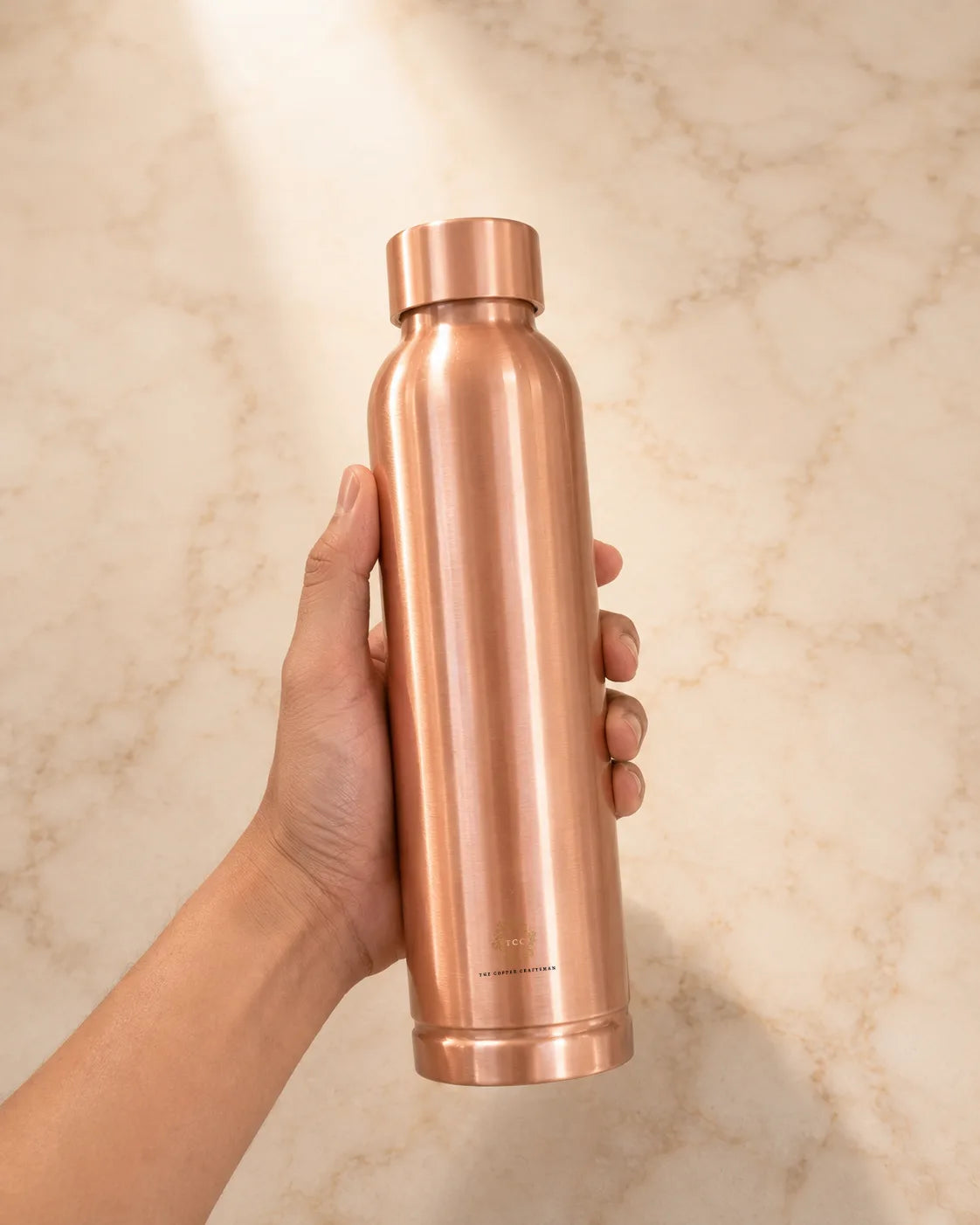 A hand holding a sleek, metallic rose gold copper water bottle against a light beige marble background.