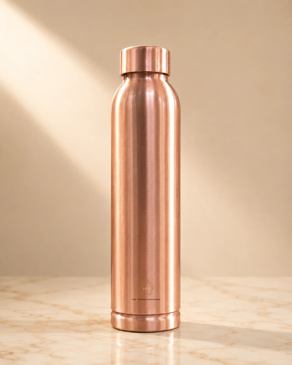 A tall, brushed copper water bottle (plain) with a matching screw-on lid, standing on a light marble surface against a warm beige background with soft diagonal light rays.