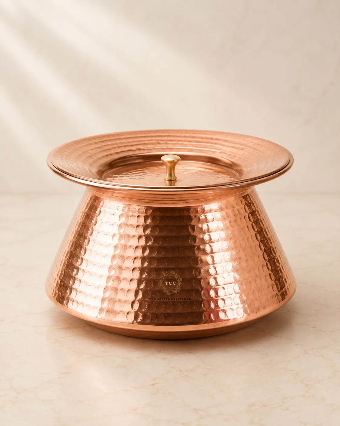 A hammered copper lidded handi with a matching copper lid and small gold-toned knob, photographed on a light beige marble-like surface with soft shadowing.