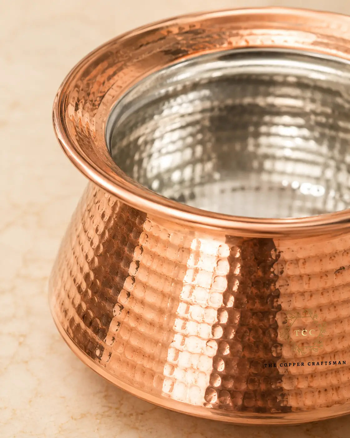 A close-up of a hammered pure-copper-handi-for-cooking with a polished silver-toned interior, resting on a light beige stone surface.