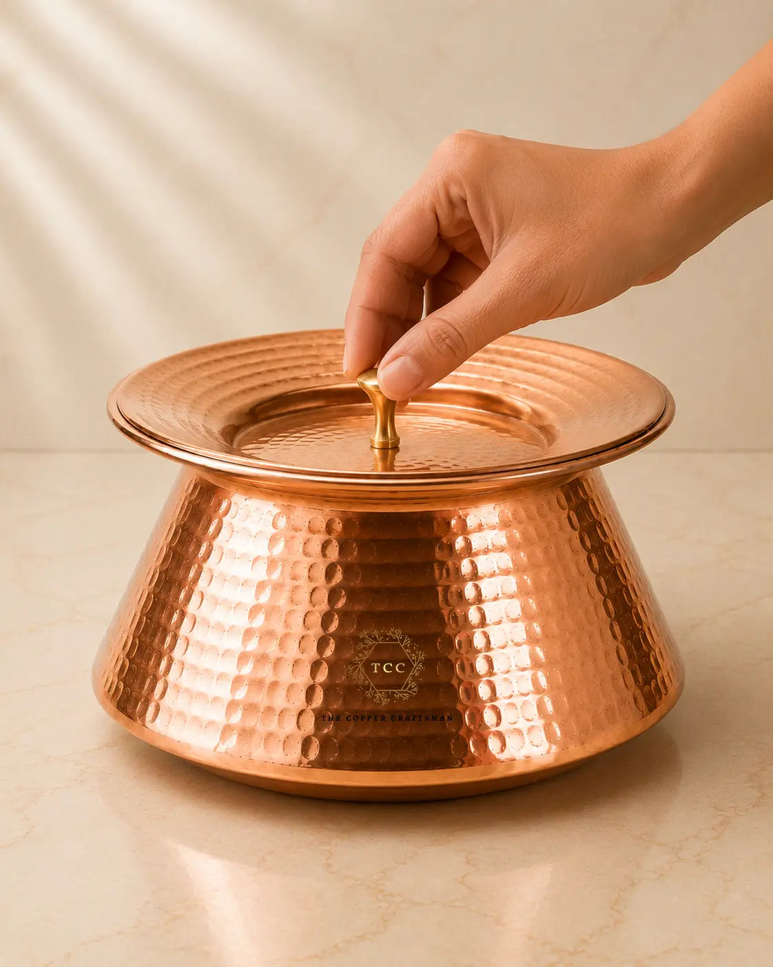 A hammered pure-copper-handi-for-cooking with a small brass handle, shown on a light beige surface with a hand lifting the lid. The product has a visible 'ICC' logo on the front.